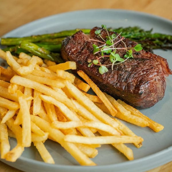 Grilled steak topped with microgreens, served with golden french fries and green asparagus on a gray plate.