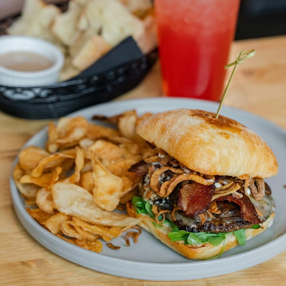 A gourmet burger with bacon, crispy onions, and arugula, served with potato chips and a red drink.
