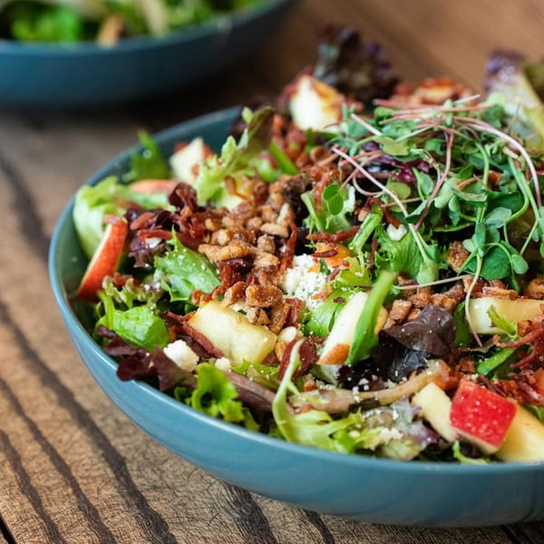 A vibrant Ednas Market & Grille salad with greens, apples, bacon, and microgreens in a blue bowl.