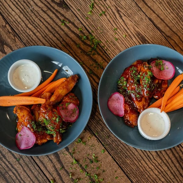 Two plates of Ednas Market & Grille's New American chicken wings with carrots, radishes, and ranch.