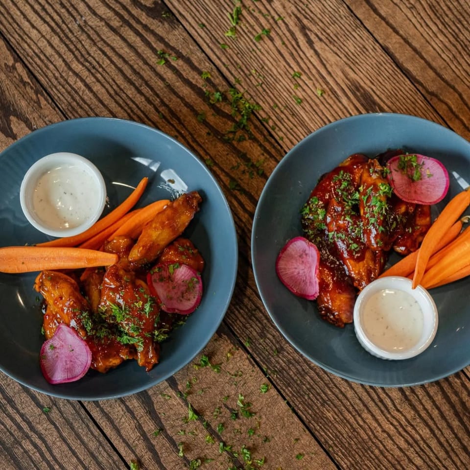 Two plates of Ednas Market & Grille's New American chicken wings with carrots, radishes, and ranch.