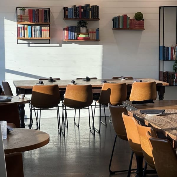 Sunlight streams into Ednas Market & Grille, illuminating tables with brown chairs and wall-mounted bookshelves.