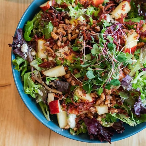 Close-up of a vibrant salad with mixed greens, apples, bacon, nuts, and microgreens in a blue bowl.