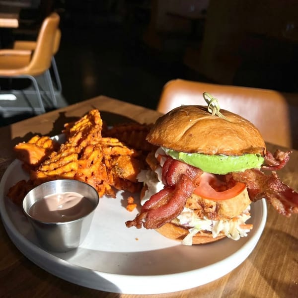 A crispy chicken sandwich with bacon, avocado, tomato, and coleslaw, served with waffle fries and dipping sauce.